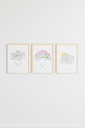 Set of three pastel cloud rainbow and sun wall art prints in wood frames on a blue wall in a children's playroom